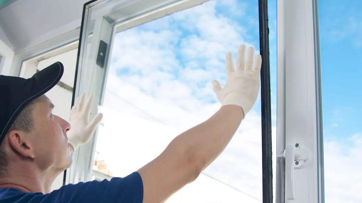Un vitrier portant des gants ajuste une fenêtre ouverte avec vue sur un ciel bleu nuageux