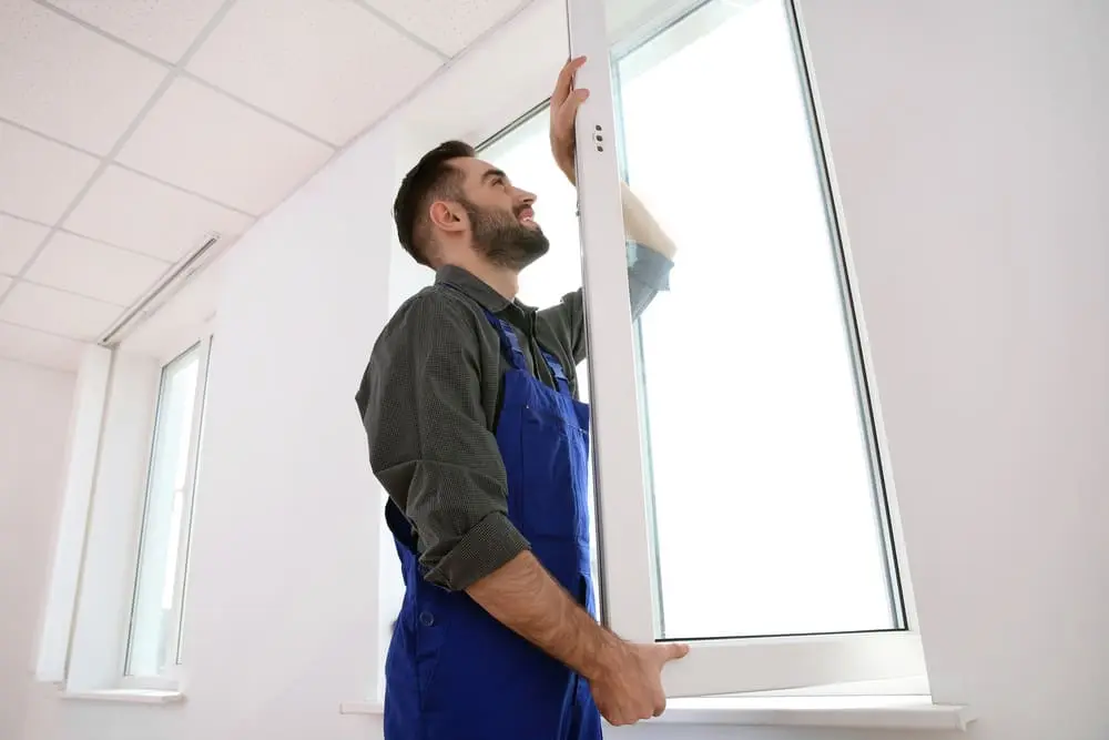 Un serrurier vitrier en salopette installe une fenêtre blanche dans une pièce.